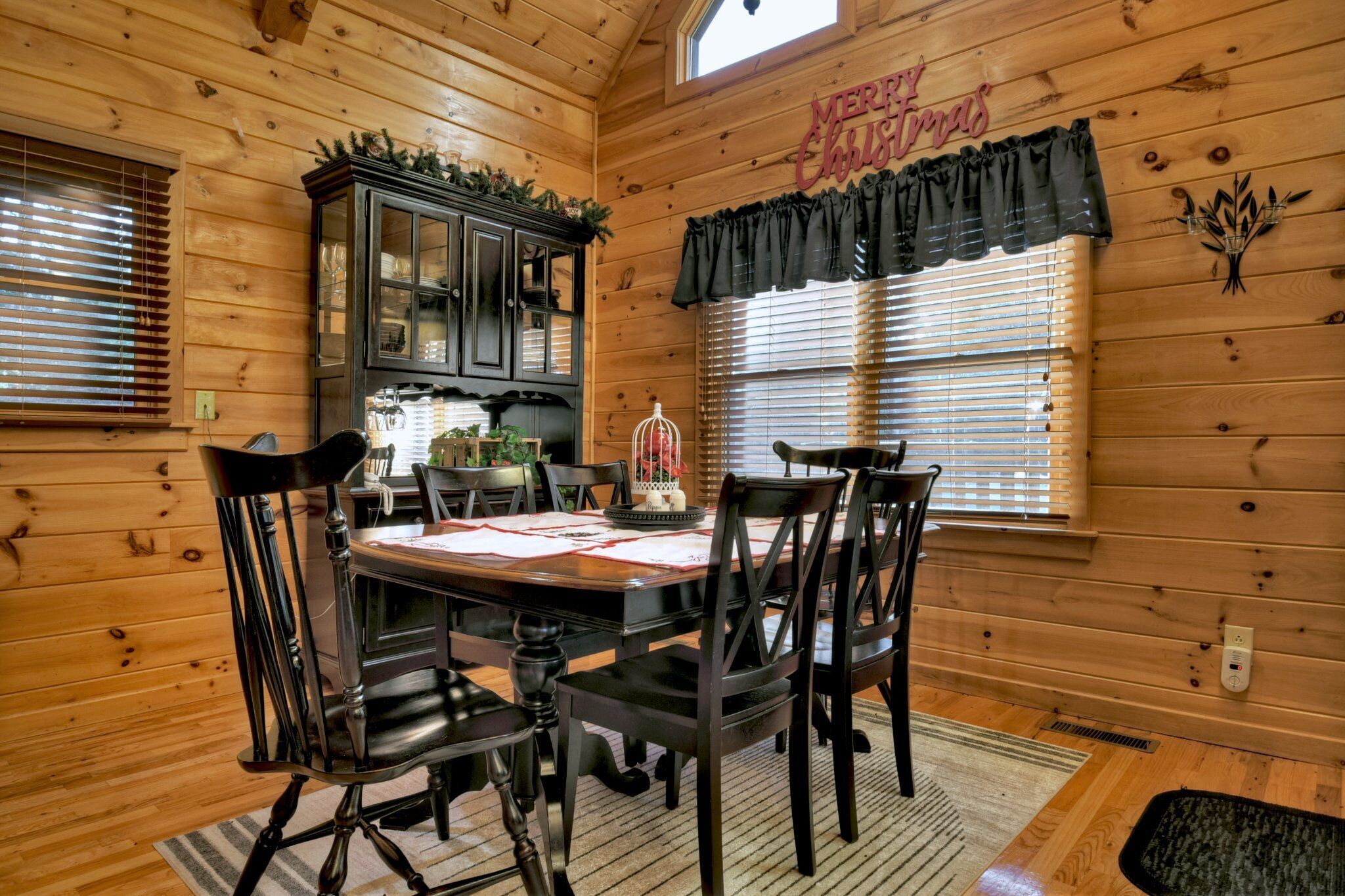 A wooden cabin interior with a dining table set for four, surrounded by chairs. A cabinet with glass doors is visible, and a 'Merry Christmas' sign hangs on the wall.