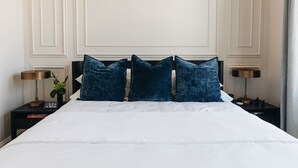 A neatly made bed with white linens and three dark blue pillows. There are two bedside tables with lamps and a clock on one of them. The room has a classic design with paneling on the walls. - The Nook at The Purple Martin is a two bedroom one bath unit in Norton Commons (Prospect)