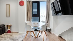 A modern dining area with a round table, white chairs, a red clock, and a large window with curtains.