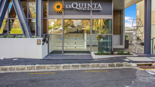 The exterior of La Quinta by Wyndham, featuring a modern design with a glass entrance, visible parking area, and a staircase leading to the entrance.