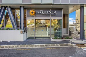 Exterior do La Quinta by Wyndham, com design moderno, entrada de vidro, área de estacionamento visível e escadaria que leva à entrada.