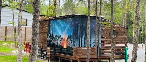 Une cabane rustique, ornée d’une fresque représentant un incendie de forêt sur l’un de ses murs, et entourée d’arbres.