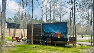 Un área de estar al aire libre de estilo rústico, con una mesa y sillas de madera, una estructura metálica y un mural grande que muestra una escena de bosque con una fogata.