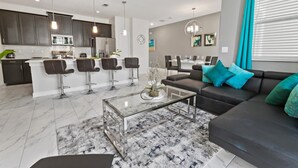 A modern living room with a glass coffee table, a dark sofa with teal cushions, and a kitchen area with dark cabinets and stainless steel appliances. - Championsgate Resort 4 Bedroom Townhome with Pool (Davenport)