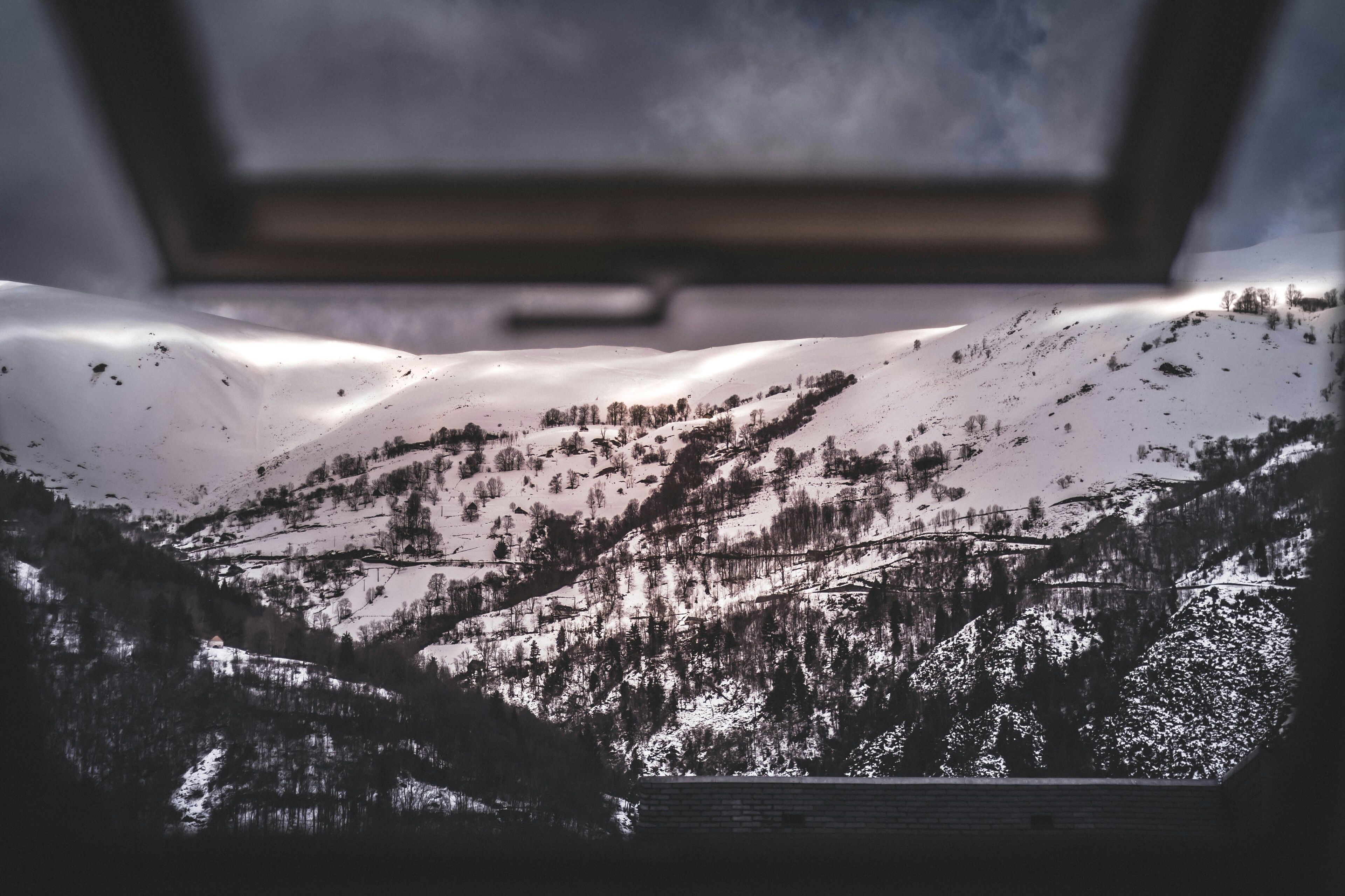A snowy mountain landscape through a window with a view of a valley and trees.