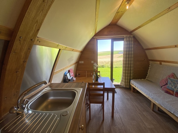 Une pièce triangulaire, chaleureuse et confortable, avec un intérieur en bois, un lavabo, une table avec des chaises et un lit donnant sur la campagne.