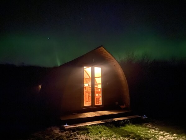 A small, dark wooden cabin with a single illuminated window, surrounded by a grassy area. - Secluded Cabin on the Banffshire Coast (Crovie)