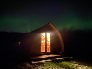 Une petite cabane en bois sombre, avec une seule fenêtre éclairée, entourée d’une zone herbeuse.