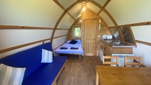 A cozy, triangular tent-style room with a bed, a blue sofa, a wooden table, and a kitchenette. - Secluded Cabin on the Banffshire Coast (Crovie)