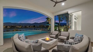 A modern outdoor seating area with a fire pit, overlooking a swimming pool and a scenic view of mountains and a lake at dusk. - Desert Flower - Contemporary villa at PGA West (La Quinta)