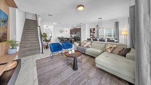 A spacious living area with a sofa, two blue chairs, a coffee table, and a staircase leading to a kitchen. - 1010 SS - Modern Comfort Oasis (Davenport)
