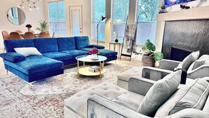 Una sala de estar moderna con un sofá seccional azul, un sillón gris y una mesa de centro. Hay una chimenea, un televisor y un ventanal con persianas.