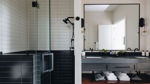 Um banheiro moderno com box de vidro, área de chuveiro com azulejos pretos e toucador com um espelho grande, pia e espaço para toalhas.