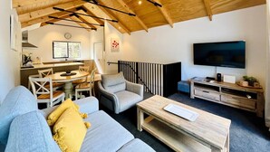 A cozy living room with a wooden ceiling, a dining area with a round table, a grey armchair, a sofa with yellow cushions, a TV on a wooden stand, and a staircase with black railings. - Queenstown City •Walk to Town •Free Parking •A/C •Balcony •Mountain Views (Queenstown)