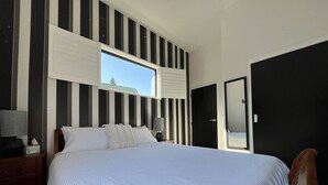 Ein Hotelzimmer mit einem großen Bett, einem Fenster mit Jalousien, einem Nachttisch mit Lampe und einer Tür.