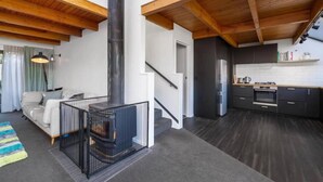 Ein modernes Interieur mit einem zentralen Kamin, einer Treppe, einer Küche mit dunklen Schränken und einem Wohnbereich mit Sofa und Teppich.