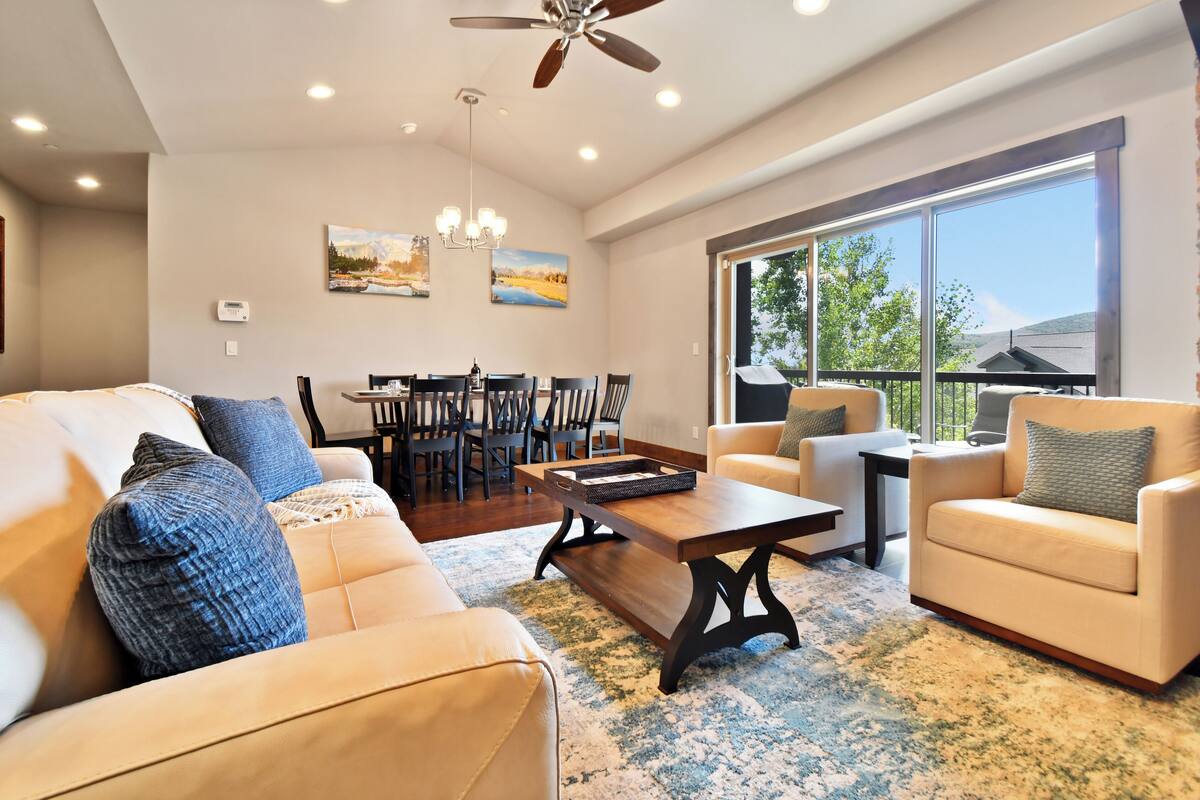 A spacious living room with a large window, a ceiling fan, and a dining area with chairs and a table.