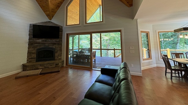 A spacious living room with a stone fireplace, large windows, and a high ceiling with wooden beams. - New Riverfront Retreat – Ultimate Privacy, Stunning Views, & Dog-friendly! (Bryson City)