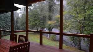 A wooden deck with a table and chairs, overlooking a forested area and a body of water. - New Riverfront Retreat – Ultimate Privacy, Stunning Views, & Dog-friendly! (Bryson City)
