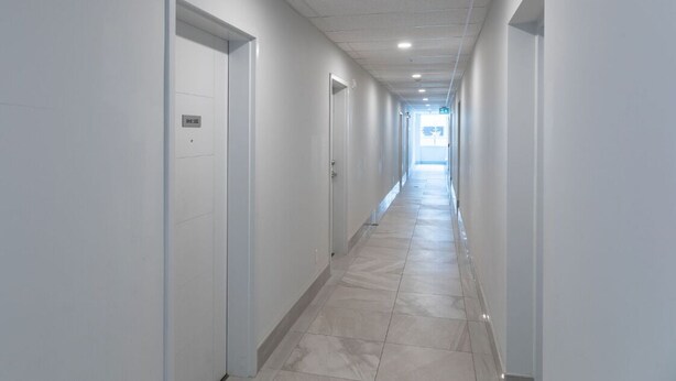 Un long couloir étroit aux murs blancs et au sol carrelé, qui mène à une sortie lumineuse au bout.