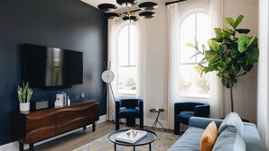 Una sala moderna con una pared oscura, un televisor de pantalla plana, un mueble de madera, dos sillones azules, un sofá, una mesa de centro y una planta grande.