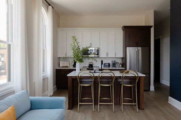 A modern kitchen with white cabinets, a stainless steel refrigerator, a microwave, and a coffee maker. There are three bar stools and a potted plant.