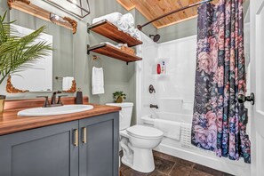 A bathroom with a wooden vanity, white sink, and a shower with floral curtains. - Cozy, stylish, and surrounded by nature; Sojourn Hideaway is the perfect getaway (Windsor)