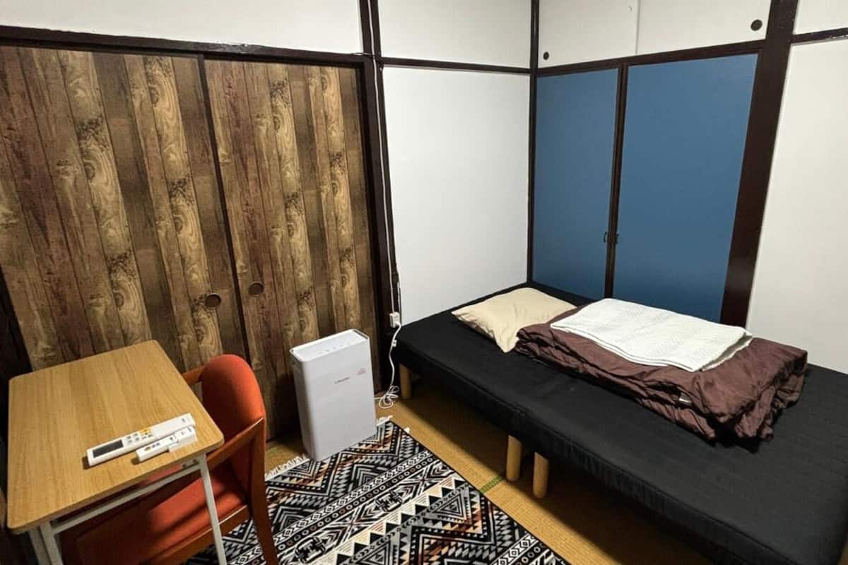 A compact room with a bed, a desk, and a chair. There is a small white appliance on the floor.
