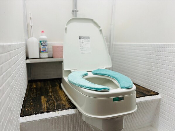 A compact bathroom with a white tiled wall, a white toilet with a teal seat cover, and a small shelf with toiletries. - SLOW HOUSE yugawara - hostel (Yugawara)