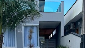 A modern two-story house with a flat roof, large windows, and a small garden with palm trees and shrubs. - Sunset House (Chácaras Mogiano Bertioga)