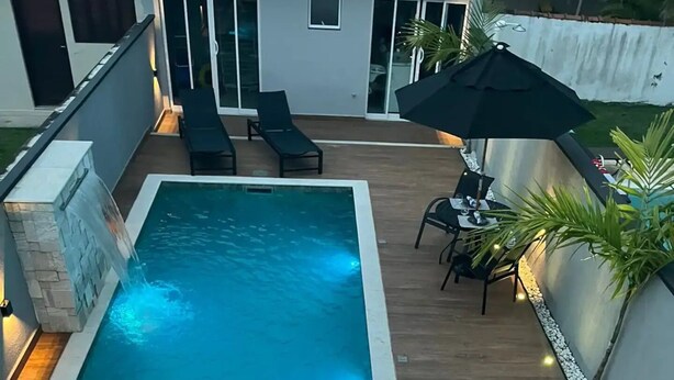 A rectangular swimming pool with a stone border, surrounded by wooden decking. There are lounge chairs and a table with chairs nearby. - Sunset House (Chácaras Mogiano Bertioga)