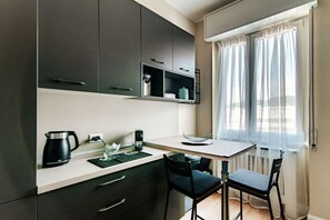Een moderne keuken met donkere kastjes, een wit aanrecht, een waterkoker, een koffiezetapparaat en een raam met lichte gordijnen.