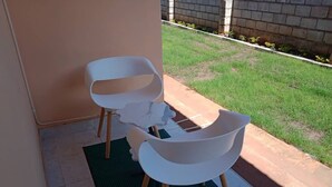 Due sedie bianche con gambe di legno su un tappetino verde, di fronte a un edificio con pavimento piastrellato.