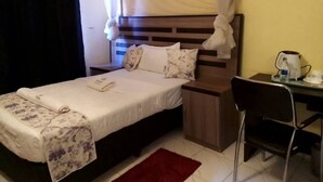 Un letto rifatto con cura, con una coperta a fiori e un baldacchino bianco. Un comodino in legno con una lampada e un piccolo tavolino si trovano accanto al letto. Un tappeto rosso è steso sul pavimento.