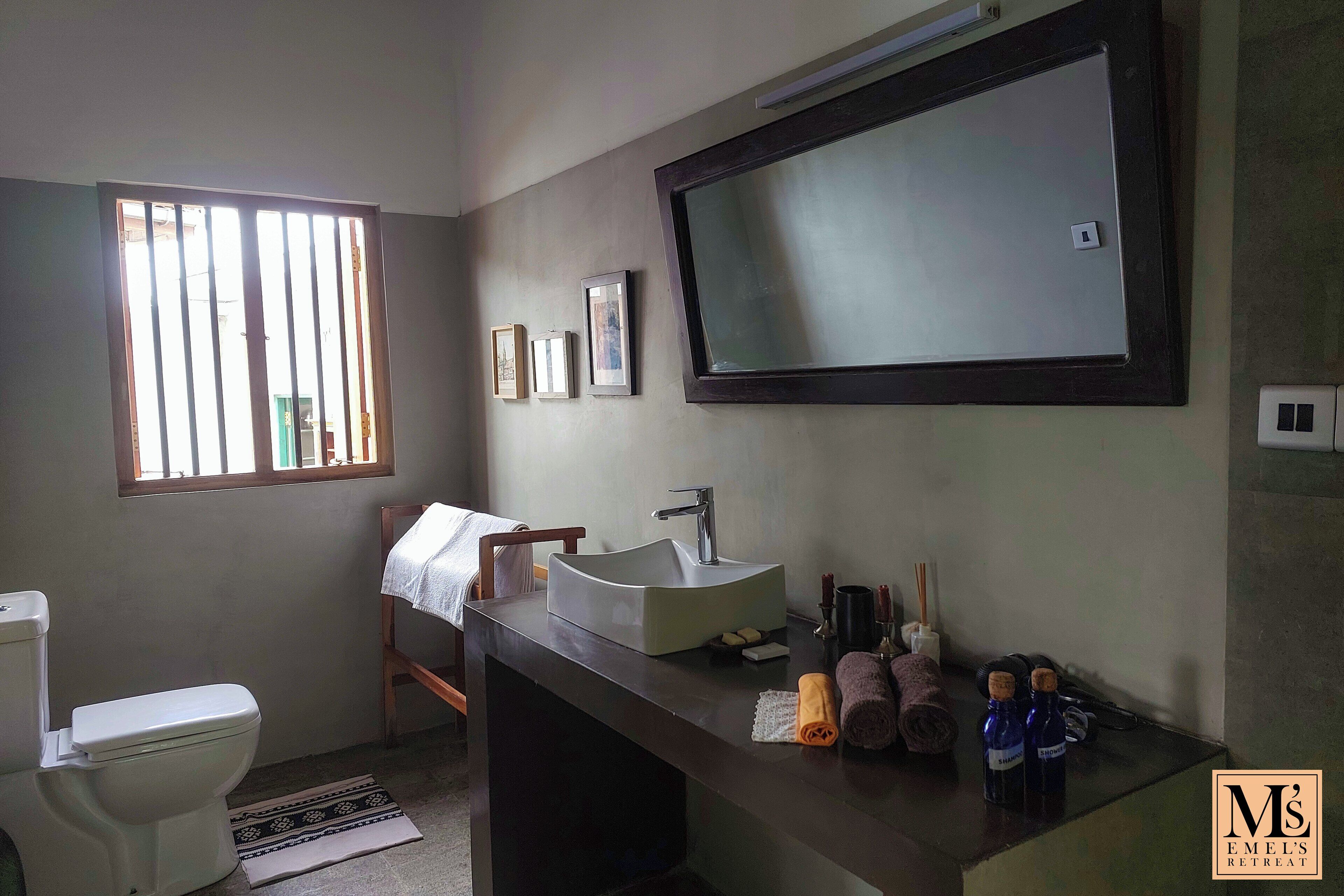 Un baño con inodoro, lavamanos y espejo. Hay cuadros en la pared, una ventana con rejas y una toalla sobre el lavamanos.