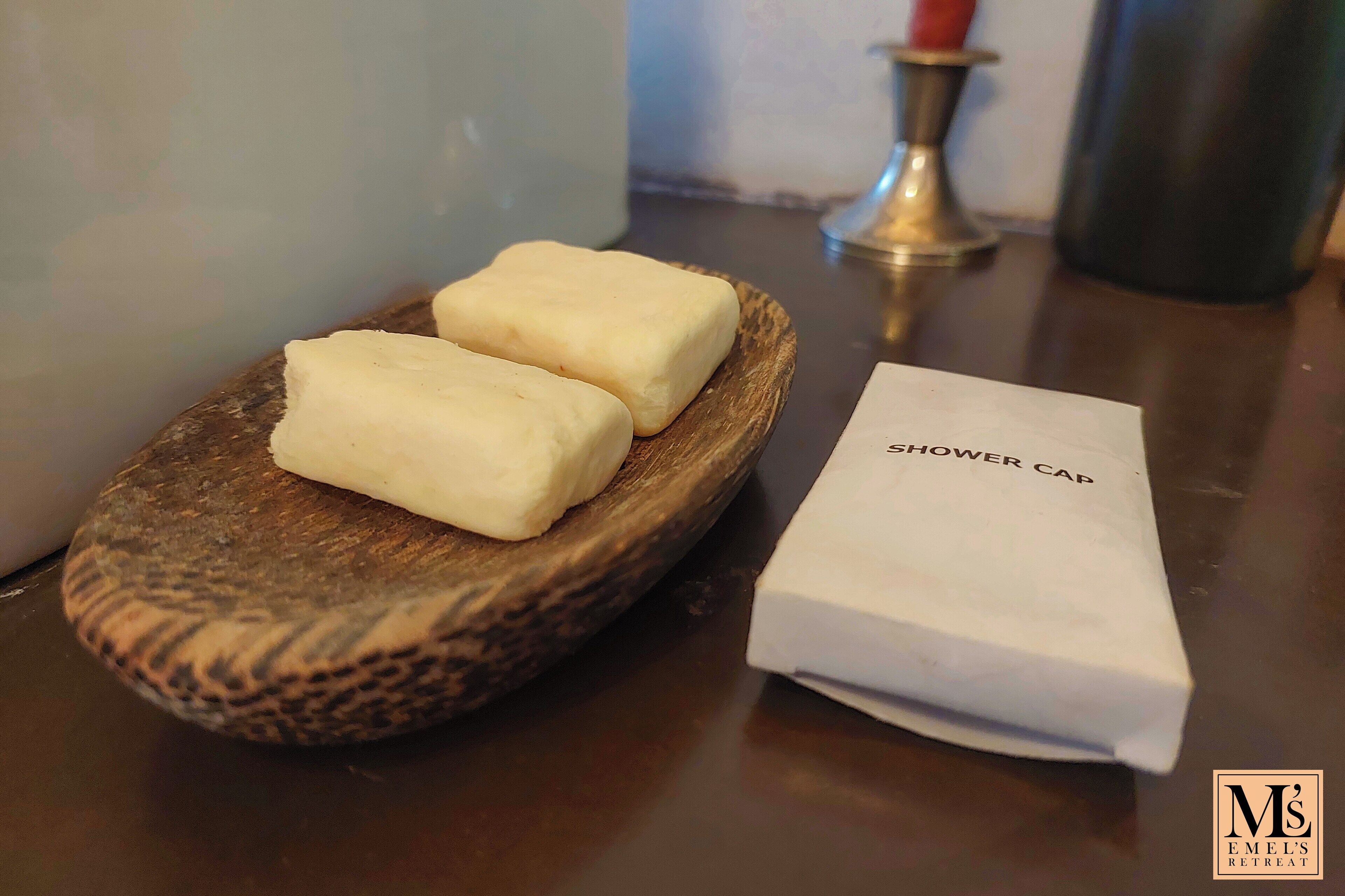 Una bandeja de madera con dos barras blancas, un papel blanco etiquetado “SHOWER CAP” y el logo  “M'S EMEL'S RETREAT” sobre una superficie oscura.