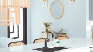 A modern kitchen with white countertops, a black sink, and a gold faucet. There are two pendant lights with clear glass shades and gold accents. A round mirror hangs on the wall above the sink, and a wicker chair is visible in the background. - Casita Canal  (Naples)