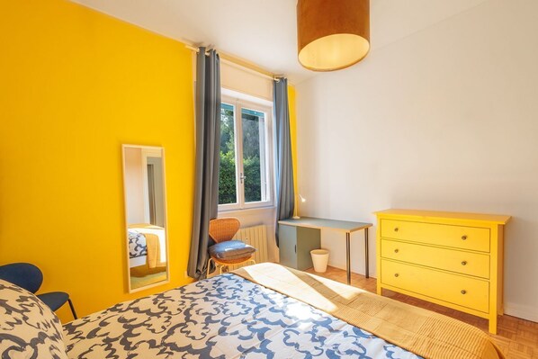 A bedroom with a bed, a desk, a chair, a dresser, a mirror, and a window with curtains.