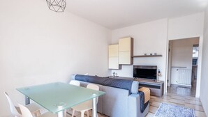 Un espace de vie moderne comprenant une table à manger en verre, des chaises blanches, un canapé gris, un téléviseur à écran plat et un coin cuisine avec des placards.