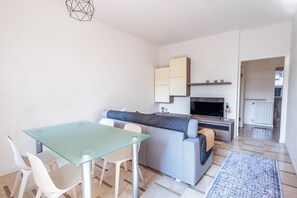A modern living space with a glass dining table, white chairs, a grey sofa, a flat-screen TV, and a kitchen area with cabinets.