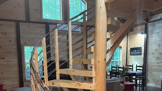Een wenteltrap van hout in een rustiek interieur met grote ramen en een gezellige zithoek.