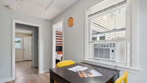 Una habitación con piso de madera, una mesa negra con sillas amarillas, una ventana con persianas y un reloj en la pared.