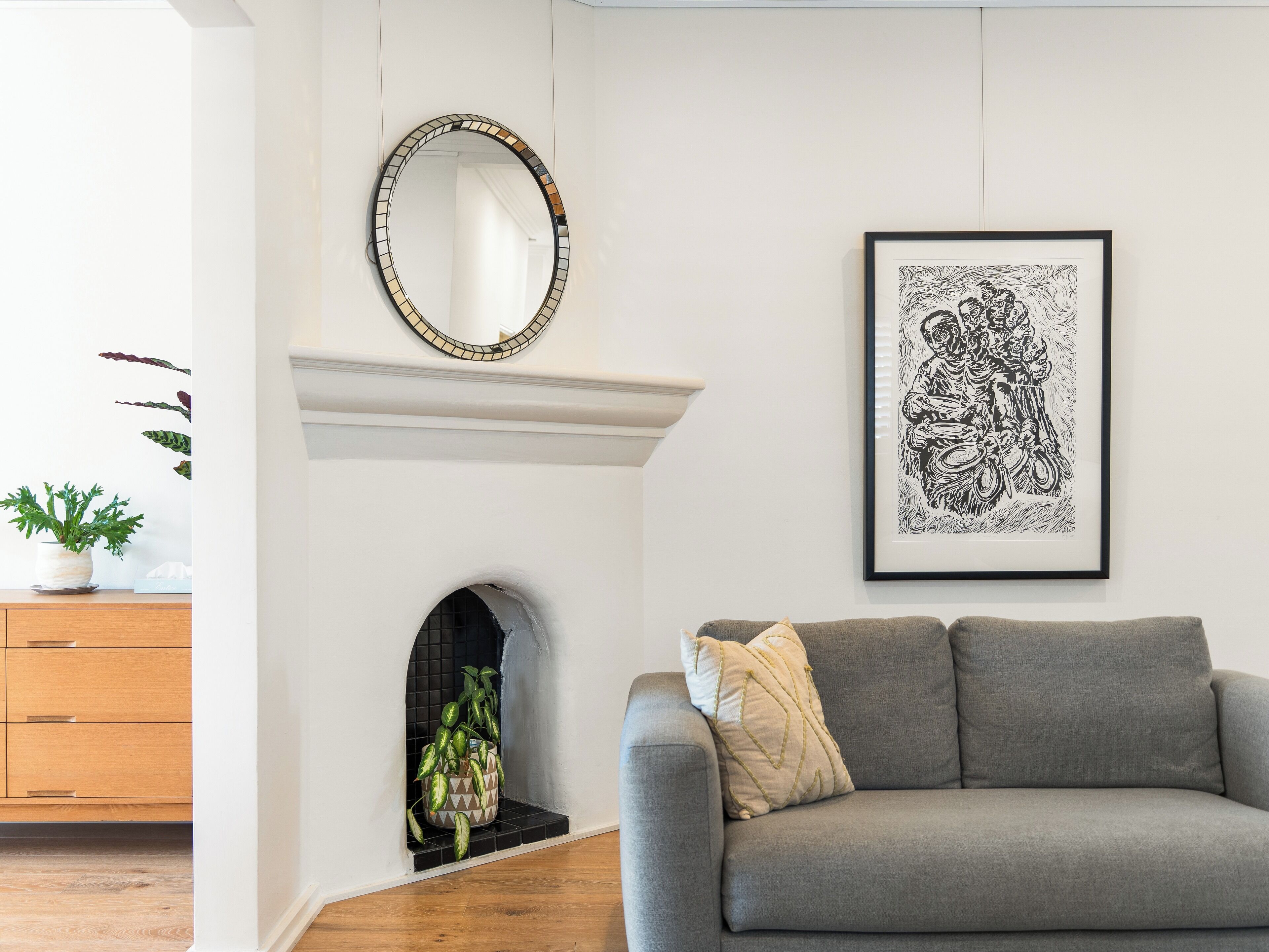 A living room with a grey sofa, a framed black and white artwork, a mirror with a mosaic border, and a potted plant on a wooden cabinet.