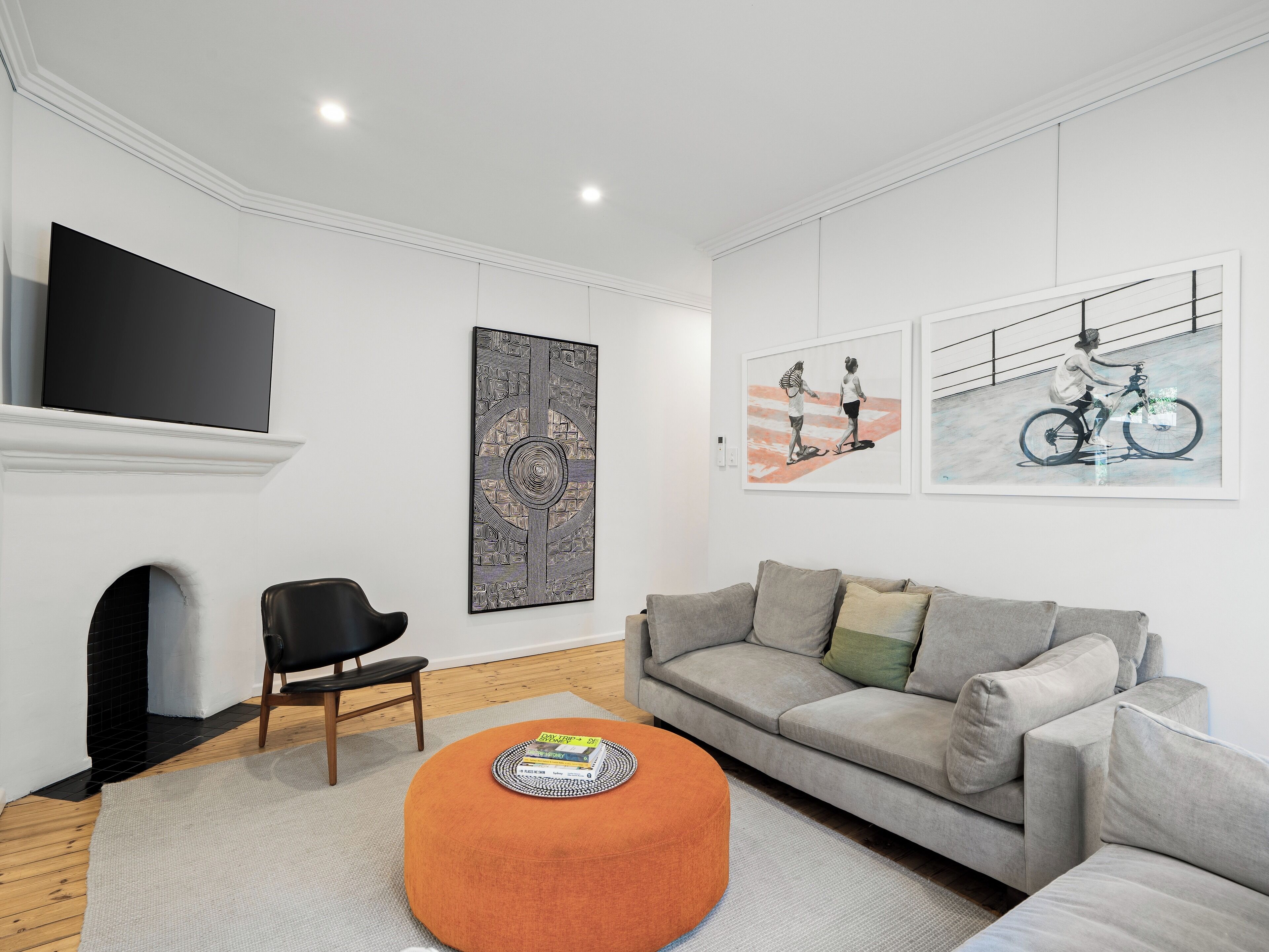 A modern living room with a grey sofa, an orange ottoman, a black chair, and a flat-screen TV mounted on the wall.