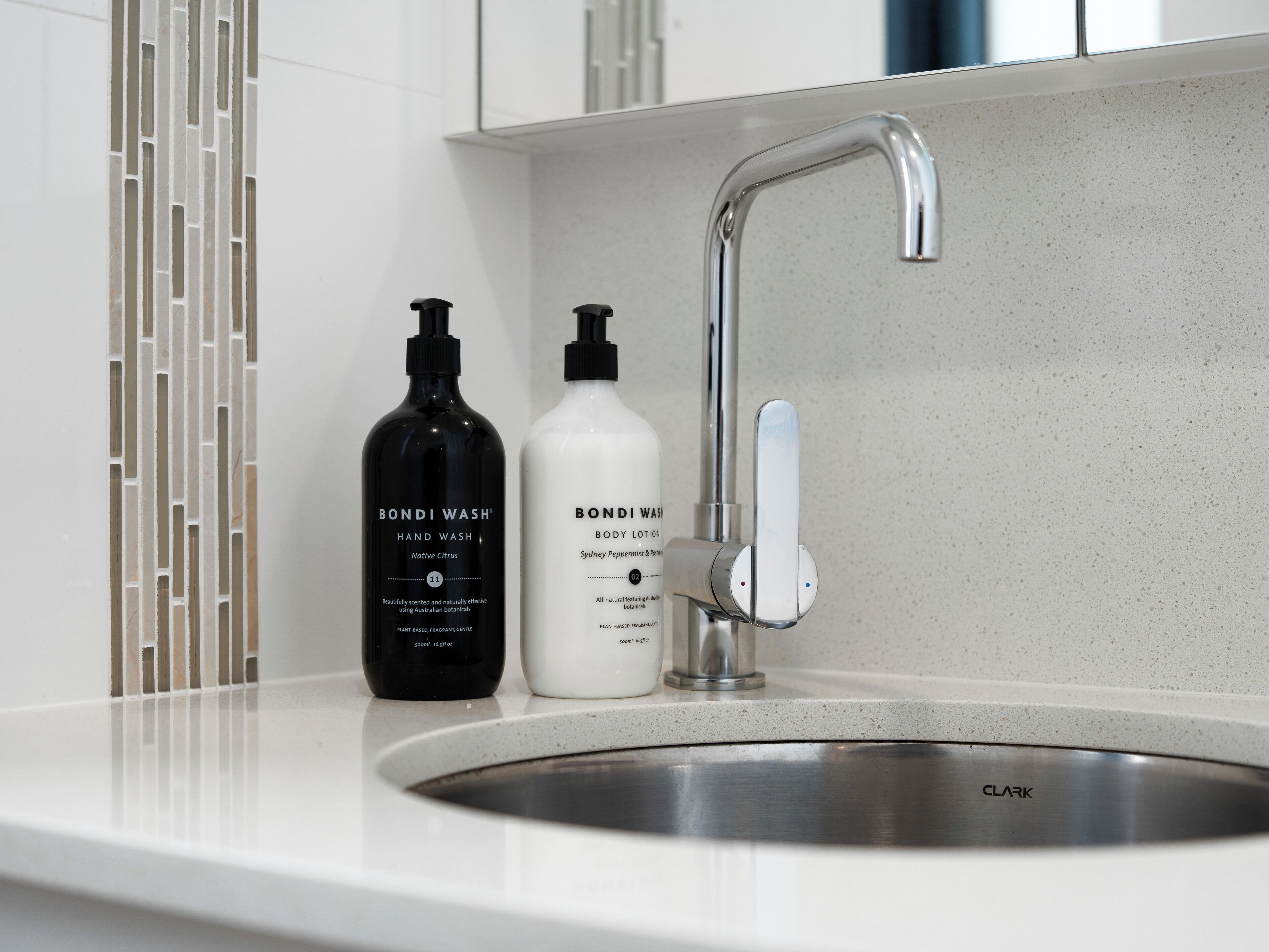 A bathroom sink with two bottles of Bondi Wash hand and body lotion, a modern faucet, and a white tiled backsplash.