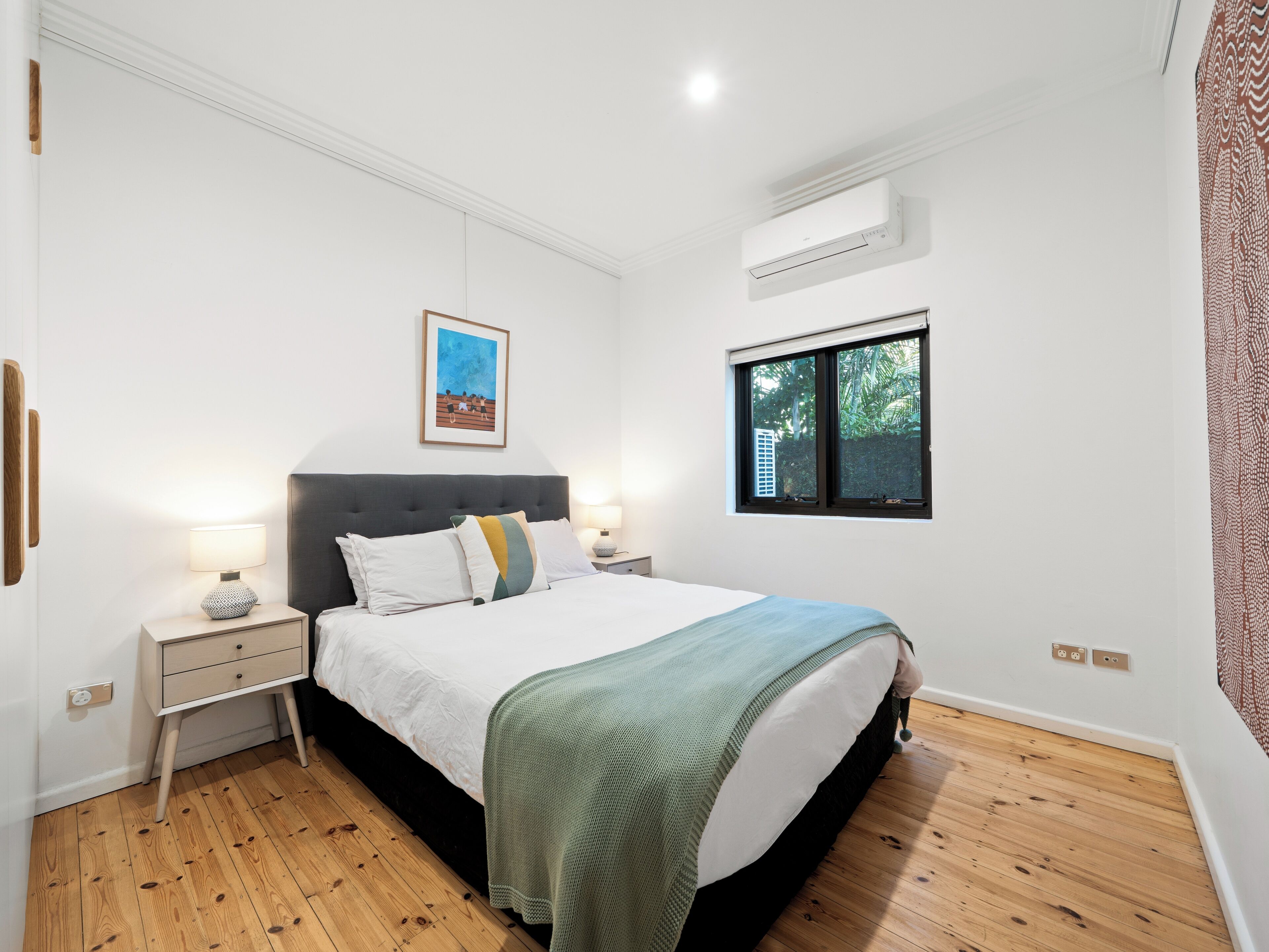 A bedroom with a bed, bedside tables, a window, an air conditioning unit, and a framed picture on the wall.