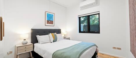 A bedroom with a bed, bedside tables, a window, an air conditioning unit, and a framed picture on the wall.