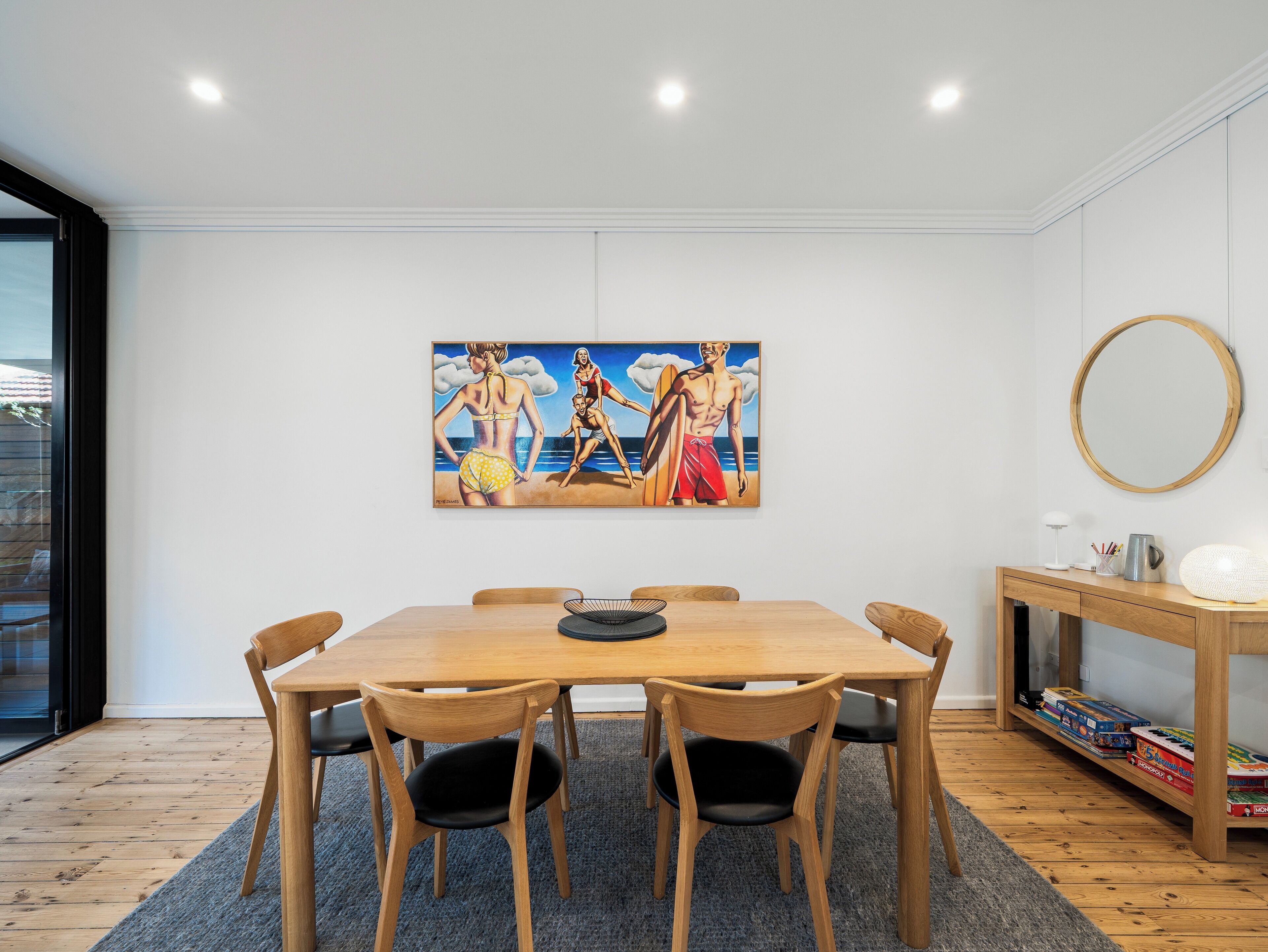 A dining area with a wooden table, six chairs, a painting of beach scenes, a round mirror, and a small side table with a lamp and a mug.