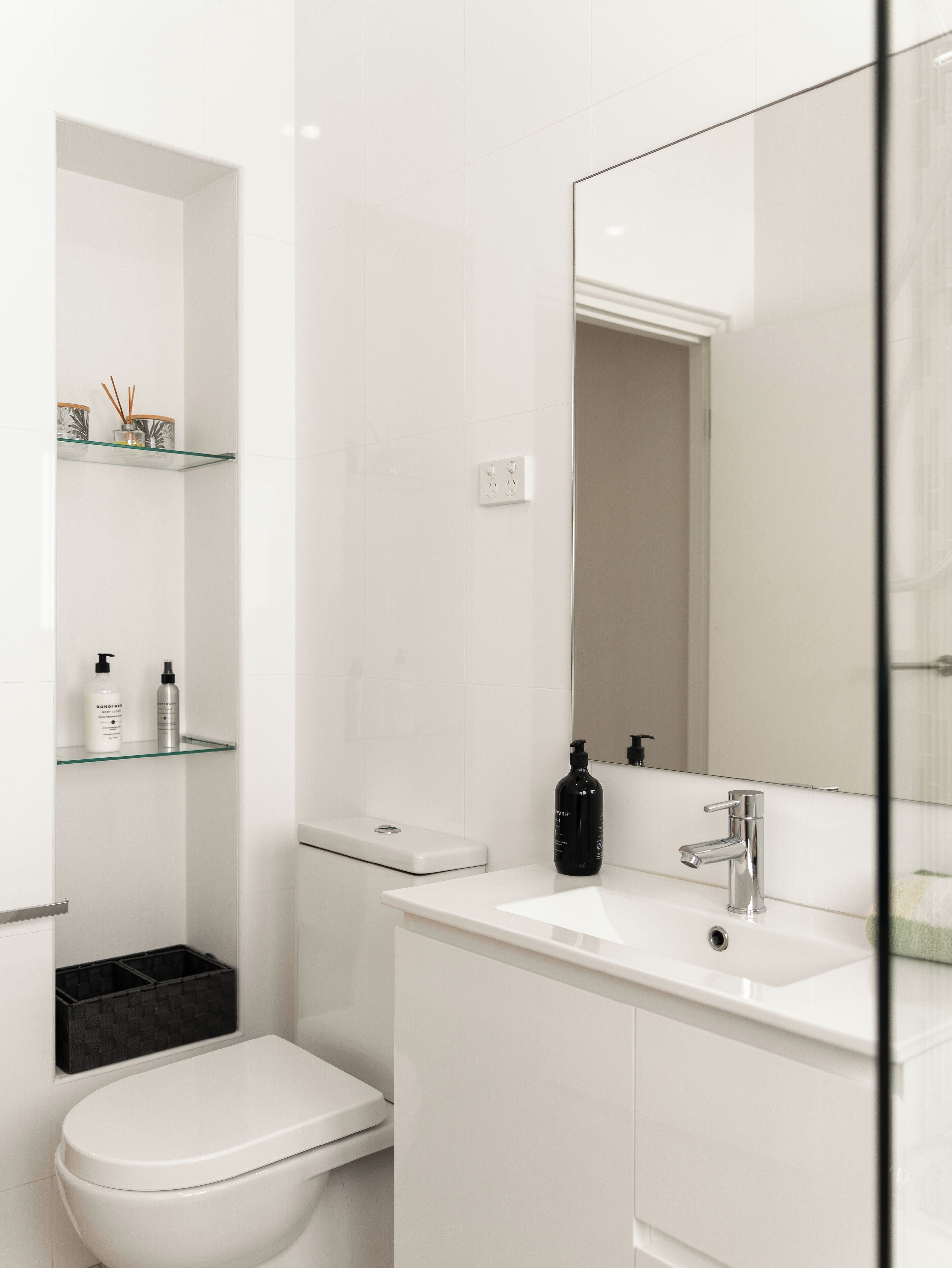 Een moderne badkamer met een wit toilet, een glazen plankje met toiletartikelen, een witte wastafel met spiegel en een zwarte zeepfles.