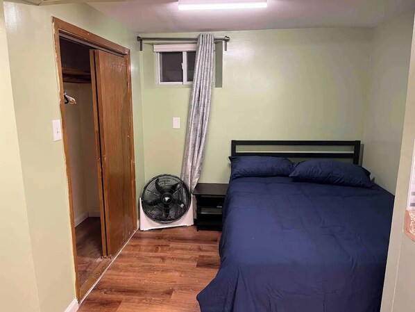 Ein Schlafzimmer mit Bett, Nachttisch, Ventilator, Fenster mit Vorhängen und Kleiderschrank.